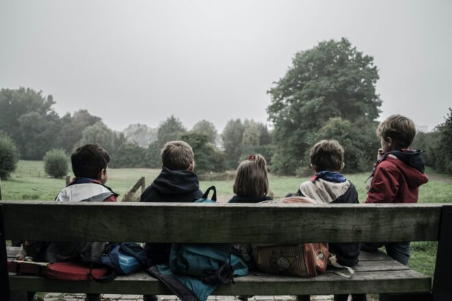 5 children sitting on a wooden bench overlooking a park with trees